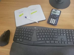 A notebook showing two steps to becoming financially independent: save and invest, alongside a calculator, keyboard, and pen on a desk.