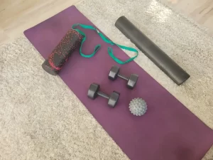 Home workout equipment on a yoga mat, including two dumbbells, two foam rollers, a green resistance strap, and a massage ball—ideal setup to start now not later on your health and wealth goals.